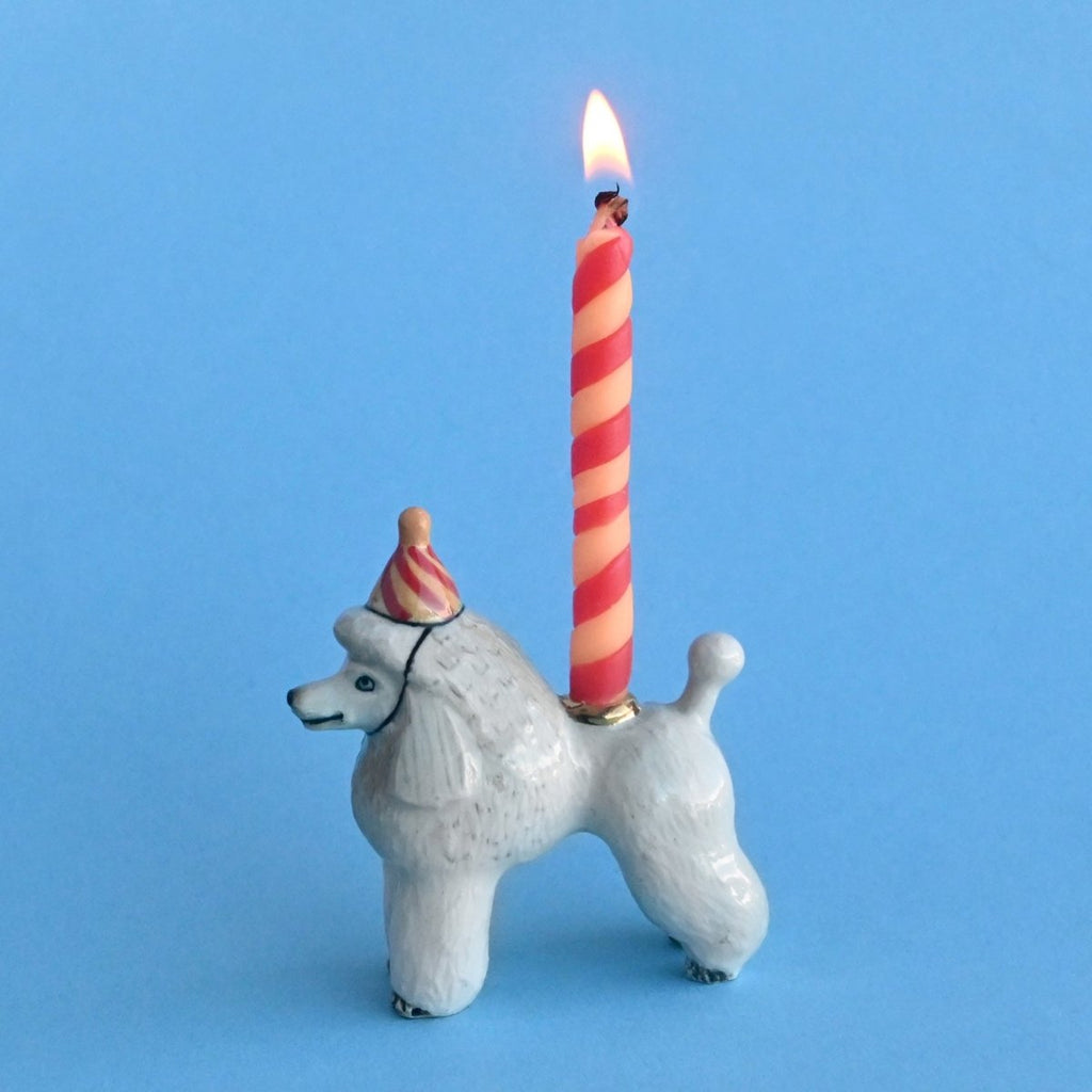 A whimsical, hand-painted Poodle Cake Topper shaped like a poodle with a birthday hat, featuring a lit pink and white striped candle on its back against a light blue background.