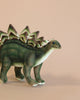 Toy model of a Stegosaurus Stuffed Animal on a pale pink background, featuring detailed textures and lifelike colors in shades of green and brown, resembling the HANSA animals collection.