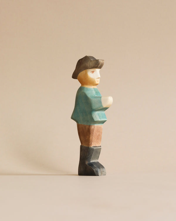 A high-quality Handmade Holzwald Farmer figurine stands against a plain, light beige background. The figurine is stylized with simple, smooth features and depicts a man wearing a hat, teal shirt, and brown pants.