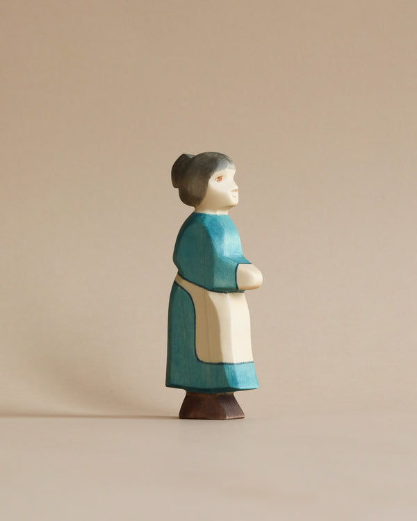 A Handmade Holzwald Grandmother figurine, painted in blue and white, standing with her arms crossed against a plain beige background. The figurine has a serene expression and simple, stylized features, ideal