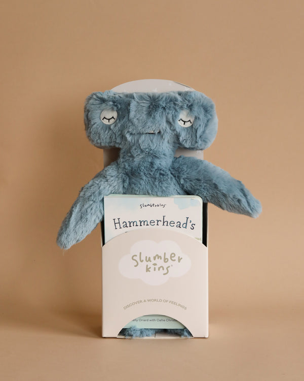 A blue plush toy resembling a hammerhead shark is partially inside a white package. The package displays the brand name "Slumberkins" and the product name "Slumberkins Hammerhead Kin + Lesson Book - Conflict Resolution." This adorable toy encourages emotional regulation and social skills development. The background is a plain beige color.