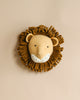 A handcrafted felt lion wall decor with a textured mane on a light beige background. The lion has a friendly face with a neutral expression and is beige with darker brown accents.