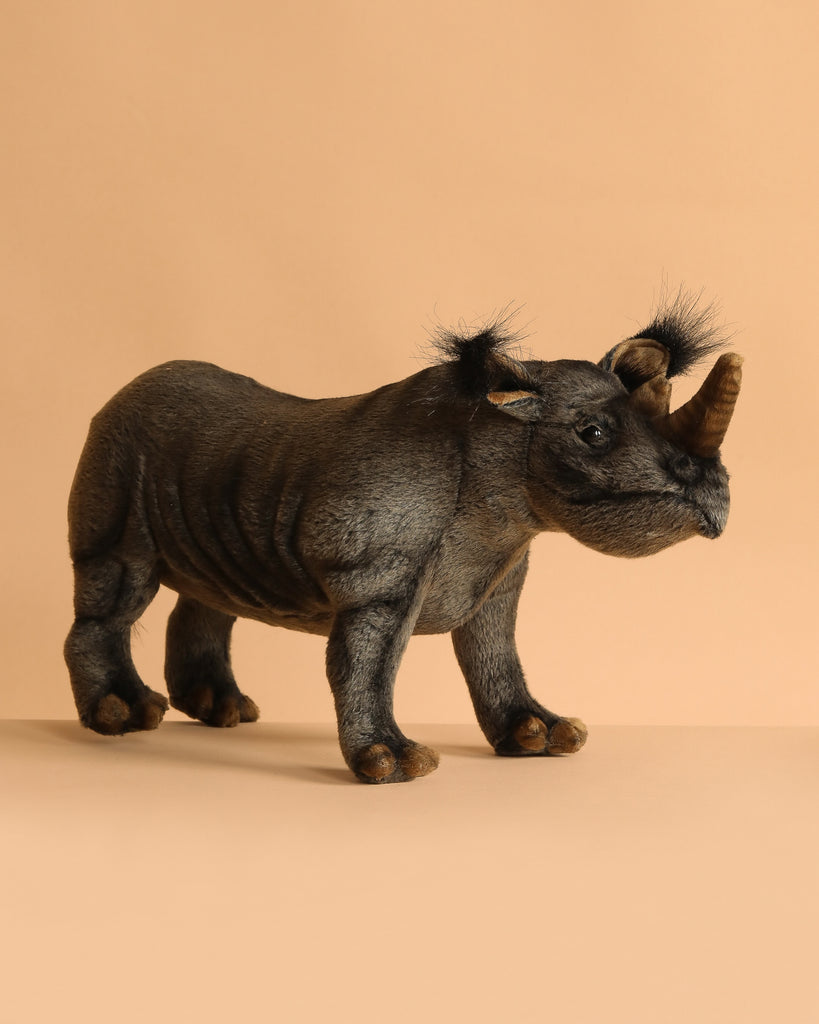 The Rhino Stuffed Animal features lifelike details with man-made fur, two horns, and black tufted ears. Hand-sewn and realistic, this plush toy stands on a beige background, perfect for showcasing quality craftsmanship.