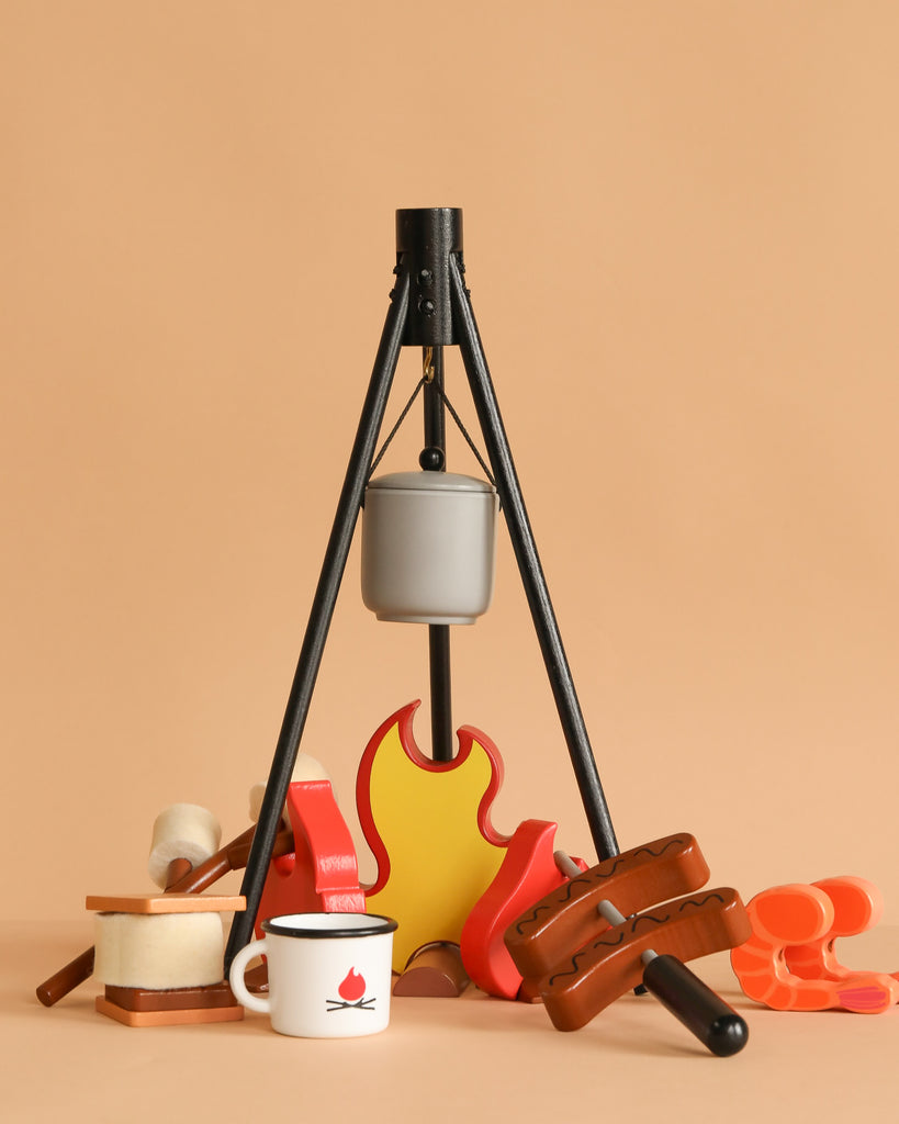 The Wooden Camp Fire Set includes a gray pot on a black tripod, toy food (bread, marshmallows, sausages), and a white mug with a red campfire logo—ideal for kids’ pretend indoor camping adventures.