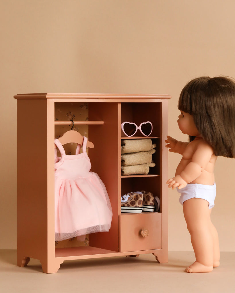 A Maileg Wardrobe, Large - Dark Powder (compatible with Minikane Dolls), stands in a light beige room, featuring a doll in white underwear, a pink dress on a hanger, bunny and teddy clothes, folded outfits, sunglasses, and a drawer.