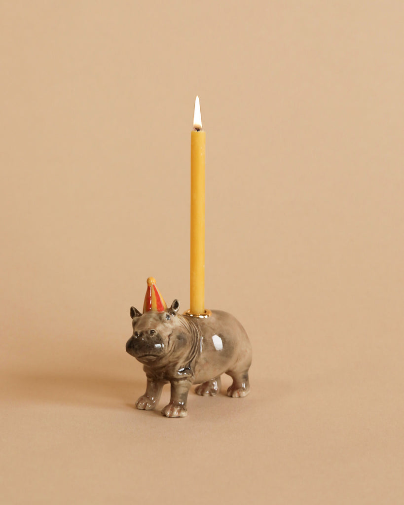 A whimsical hand-painted ceramic Hippo Cake Topper with a small red crown on its head, holding a lit yellow taper candle on its back, set against a light tan background.