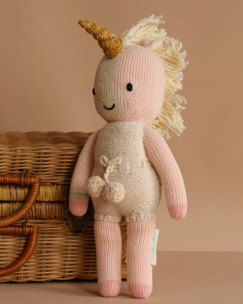 Meet Cuddle + Kind Ella the Unicorn: a hand-knit pink unicorn doll with a golden horn, cream mane, and embroidered smile, standing in front of a wicker basket on a beige background—ready to be your child’s best friend.