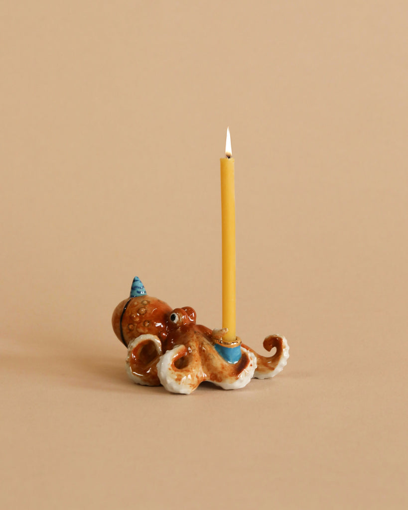 A whimsical Octopus Cake Topper shaped like an octopus, hand painted and crafted from fine porcelain, with a lit yellow candle standing upright from its center, against a soft beige background.