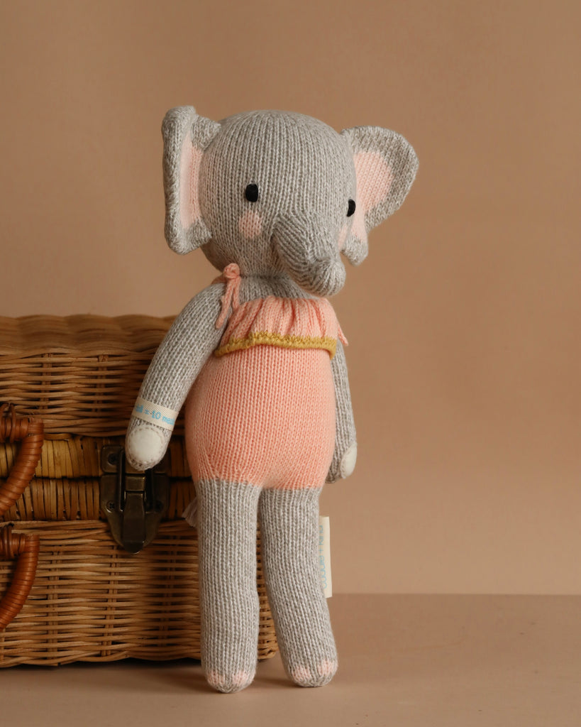 Cuddle + Kind Eloise the Elephant is a hand-knit gray elephant in a peach and pink outfit, posed by a wicker basket on beige. Each doll purchased provides 10 meals to children in need.