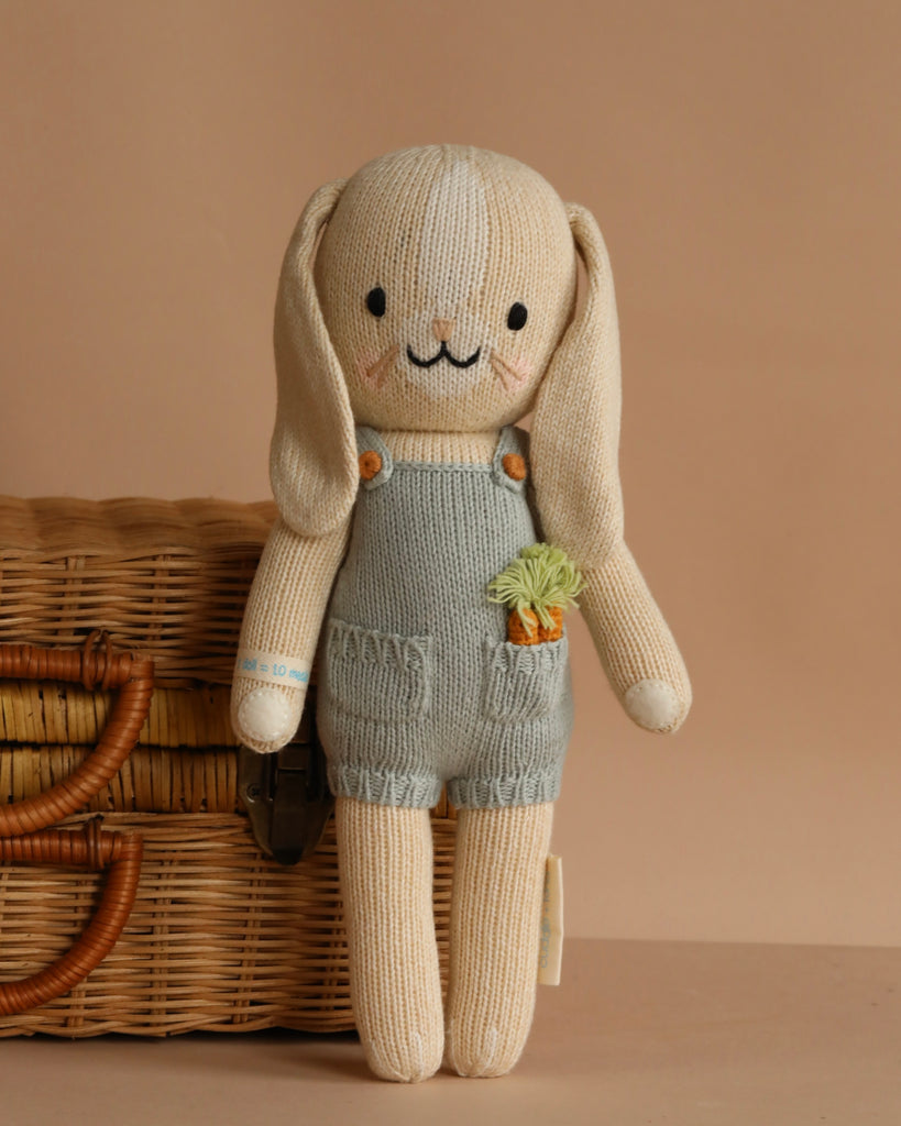 Cuddle + Kind Henry the Bunny is a hand-knit doll with long floppy ears, wearing blue overalls with a carrot detail. Made from 100% cotton yarn for softness and durability, it stands beside a woven basket against a beige background.