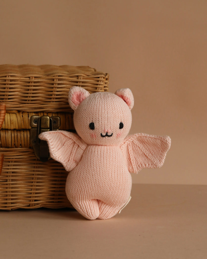 The Cuddle + Kind Baby Limited Edition Powder Pink Bat—hand-knit with bat wings, bear ears, and a smiling face—sits in front of a closed wicker basket on a beige background.