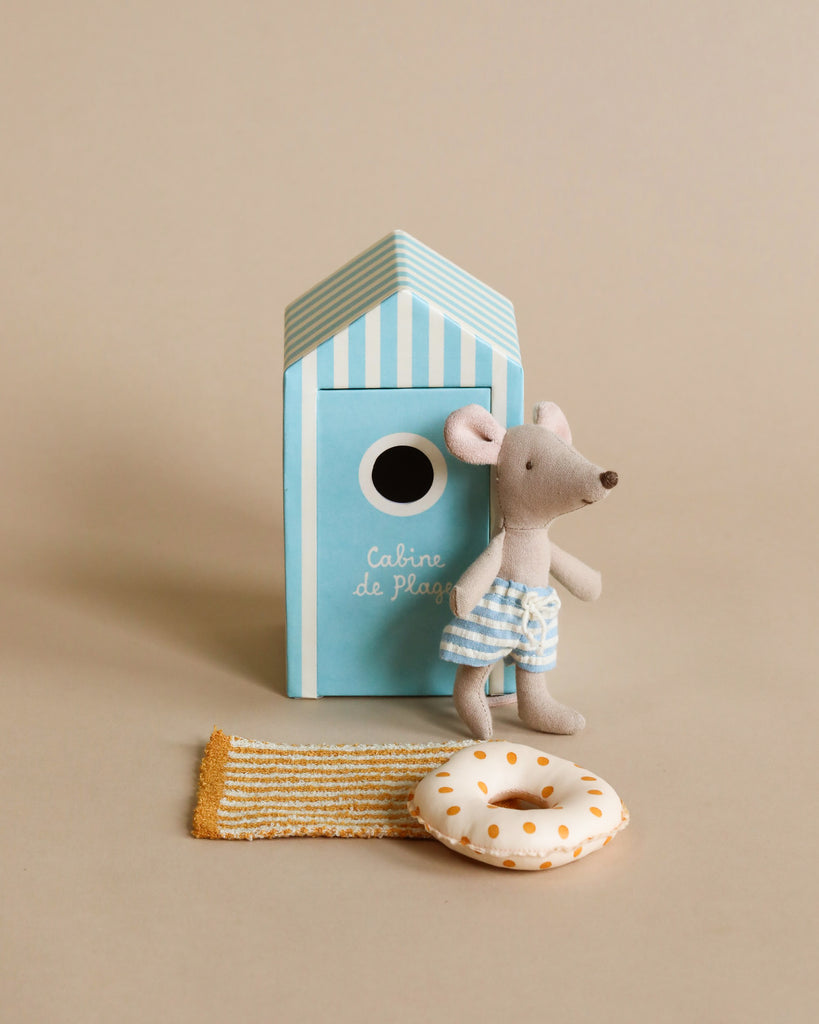 A small Maileg Beach Mouse - Little Brother in a striped blue outfit stands next to a light blue and white striped toy beach house. The scene includes vacation accessories like a miniature striped beach rug and a small white lifebuoy with orange dots. The background is a neutral beige, evoking vacation time bliss.