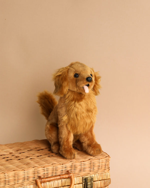 A fluffy tan realistic Golden Retriever dog stuffed toy sitting attentively on a wicker suitcase against a neutral beige background. The dog looks playful with its tongue sticking out slightly.