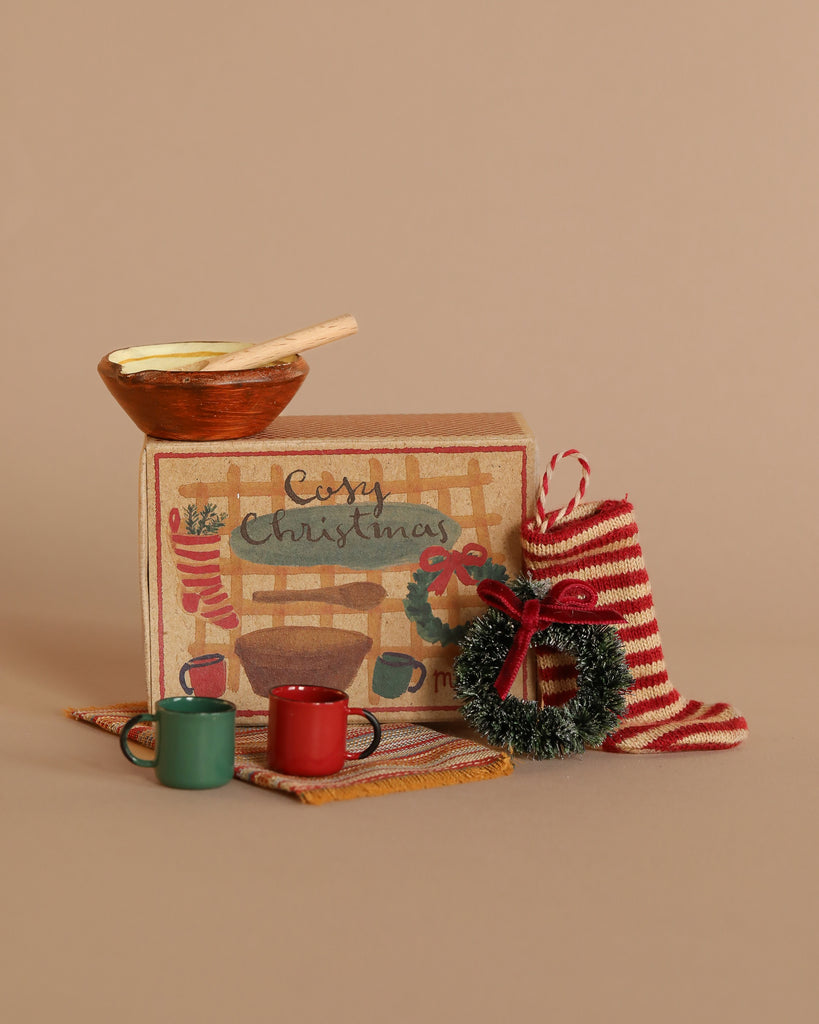 A festive scene featuring the Maileg Cozy Christmas Set with a decorative box, accessories, tiny mugs, striped stocking, mini wreath, wooden bowl with spoon, and a knit mat on a beige background.