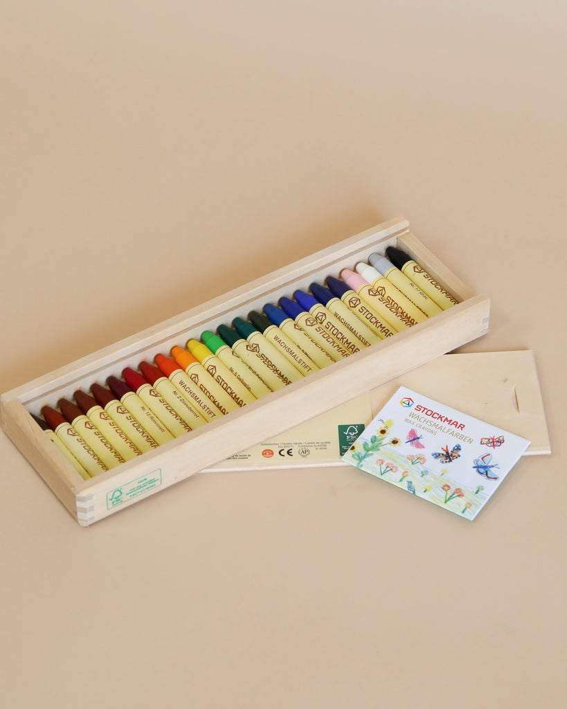 A wooden box containing rows of colorful Stockmar Wax Stick Crayons, each labeled with its color name, accompanied by a small sheet of paper with crayon drawings.