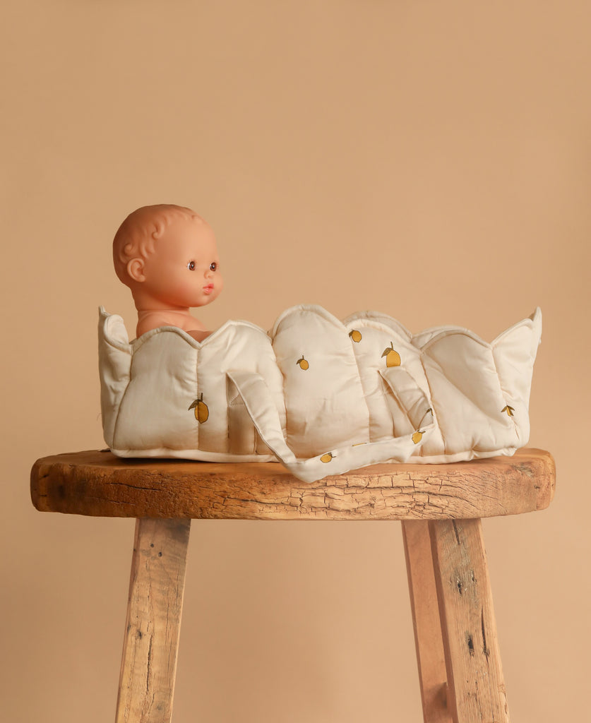 A Konges Sloejd Doll Lift - Lemon, made from soft, OEKO-TEX certified white fabric with small yellow lemon designs, holds a baby doll sitting on a rustic wooden stool against a plain beige background.