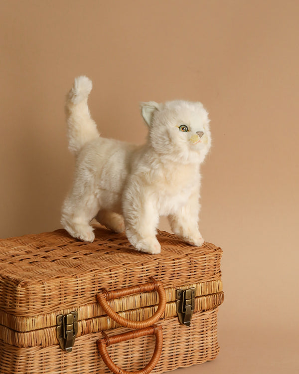 A lifelike White Cat Stuffed Animal, hand sewn by artisans, stands on a woven wicker basket with brass clasps, against a soft beige background.