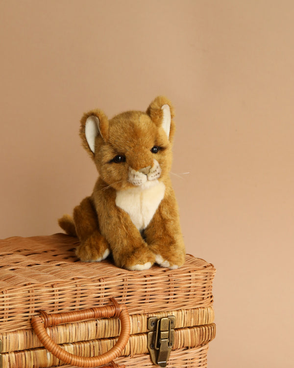 A Lion Cub Stuffed Animal, hand-sewn with lifelike features and a gentle expression, sits atop a woven wicker basket against a soft beige background.