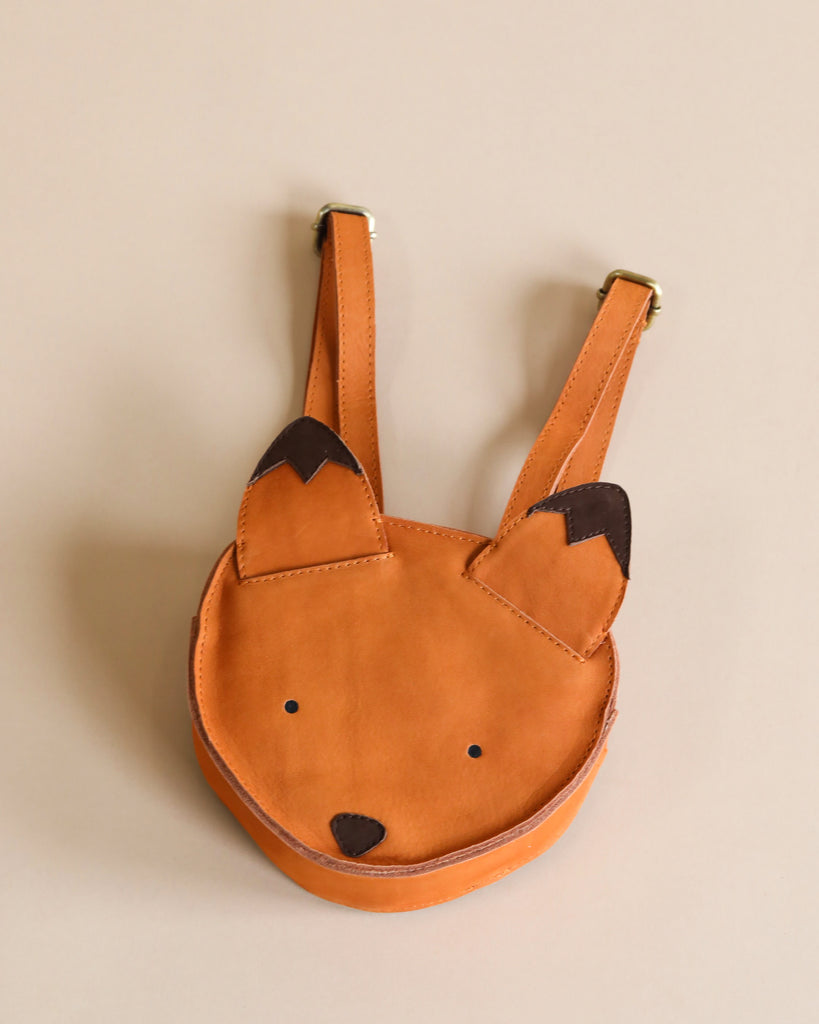 A cute Donsje Mini Leather Backpack - Fox with a tan body and dark brown ear tips, designed by Donsje, features a simple face with two black eyes and a nose, hanging on a light beige wall.