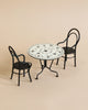 A Maileg Bistro Table With Chairs set featuring two black metal chairs with rounded backs and a small round dining table. The tabletop is white with a black decorative pattern, ideal for enjoying Sunday supper. The set resembles classic bistro-style furniture, placed on a plain beige background.