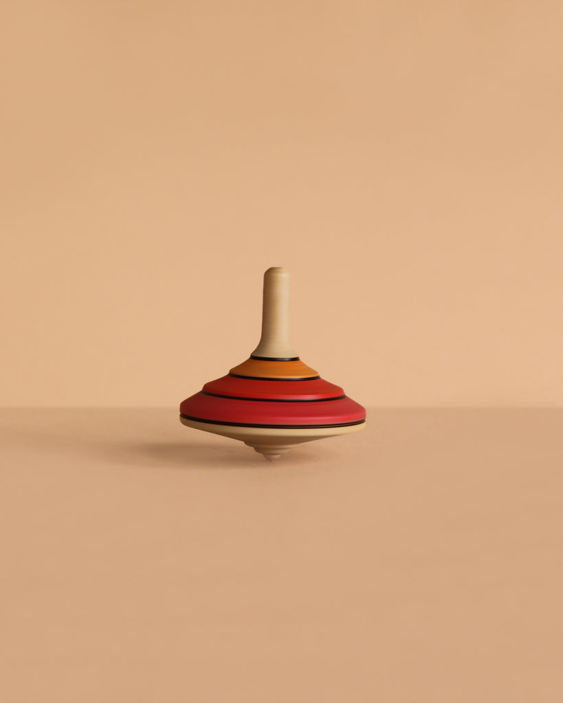 The Mader Tango Spinning Top - Red, featuring red, orange, and tan stripes and crafted from maple wood, stands upright on a smooth beige surface with a matching background.