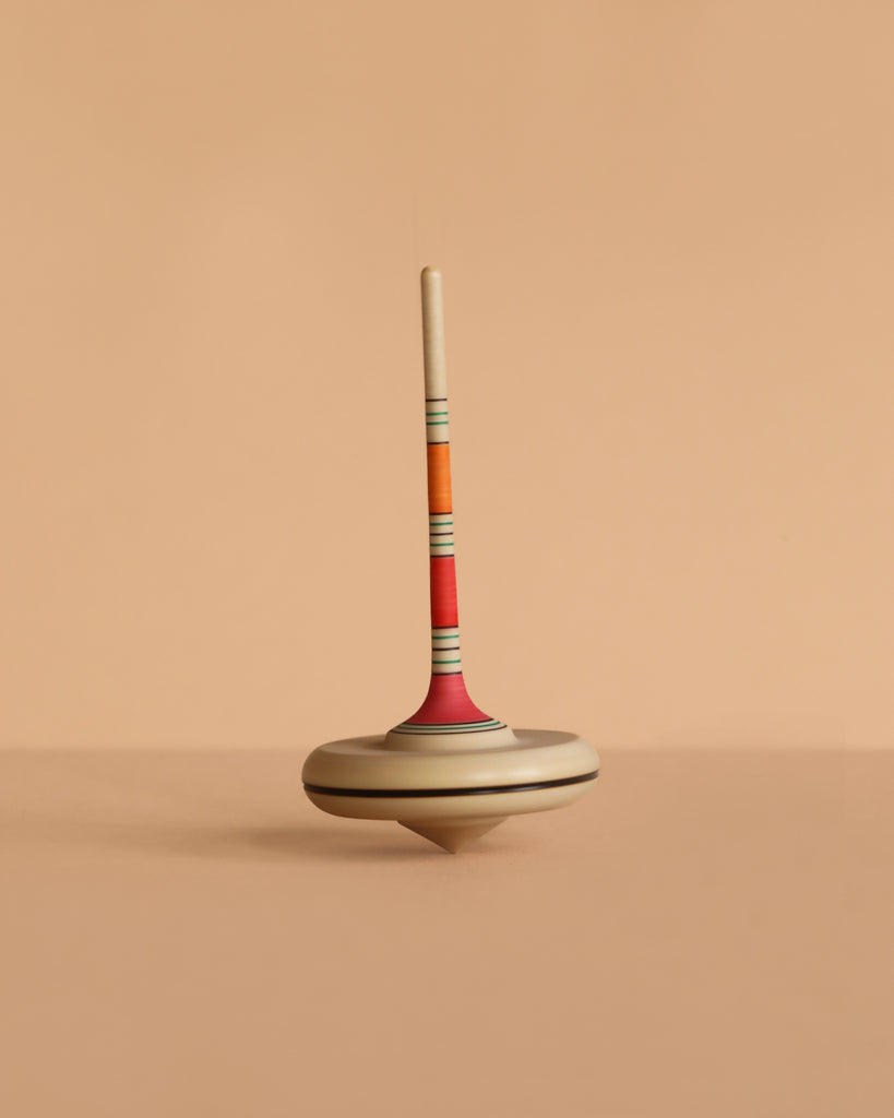 The Mader Spaghetti Spinning Top - Orange, a colorful wooden toy with a striped handle, stands upright on a beige surface with a matching beige background.