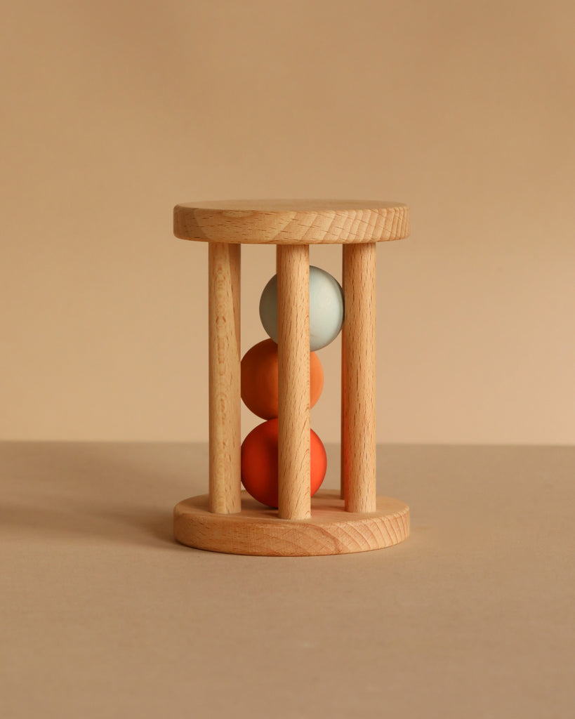 The Sabo Concept Rolling Rattle - Lagoon features three vertical rods and three colored balls—green, orange, and red—stacked inside. Set against a plain beige background, this wooden baby toy also makes a charming handmade keepsake.