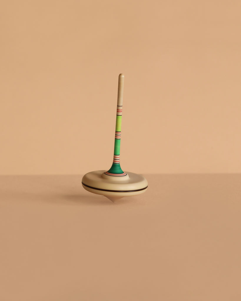 The Mader Spaghetti Spinning Top - Green, featuring a striped handle and crafted from Maple wood, stands upright on a smooth beige surface with a matching plain background.