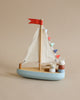 A colorful Wooden Sail Boat with fabric sails, featuring a blue base and multi-colored triangle flags. Two cute animal figures, a bear and a mouse, peek out from inside the boat.