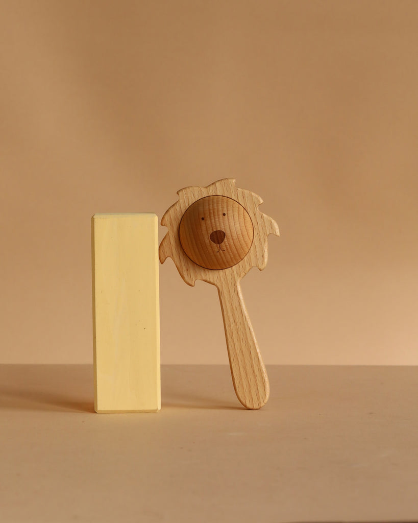The Sabo Concept Lion Rattle - Natural, featuring a carved face and mane, stands upright next to a plain yellow wooden block against a beige background, highlighting the natural charm of this baby toy.