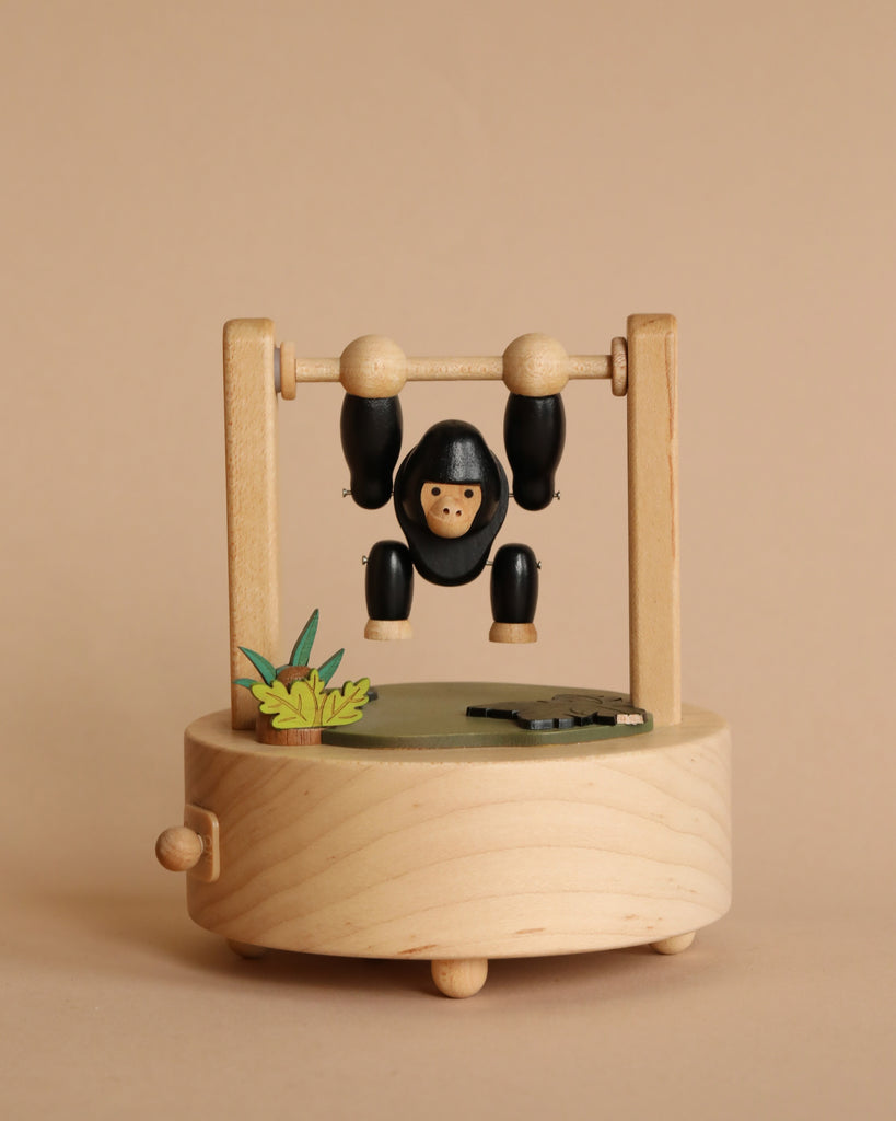 Sentence with product name:
A Gorilla Music Box from Wooderful Life, featuring a black and white monkey hanging from a horizontal bar, positioned above a round base with green leaves, all crafted from sustainably sourced wood, set against a beige background.