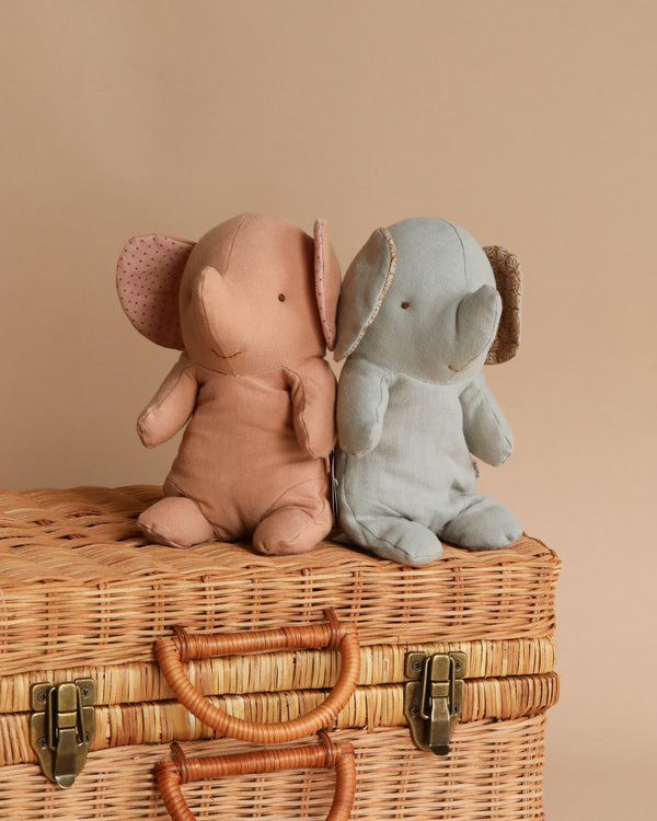 Two Maileg Small Elephant toys, one pink and one gray, crafted from the softest fabric, sitting side by side on a wicker chest against a beige background.