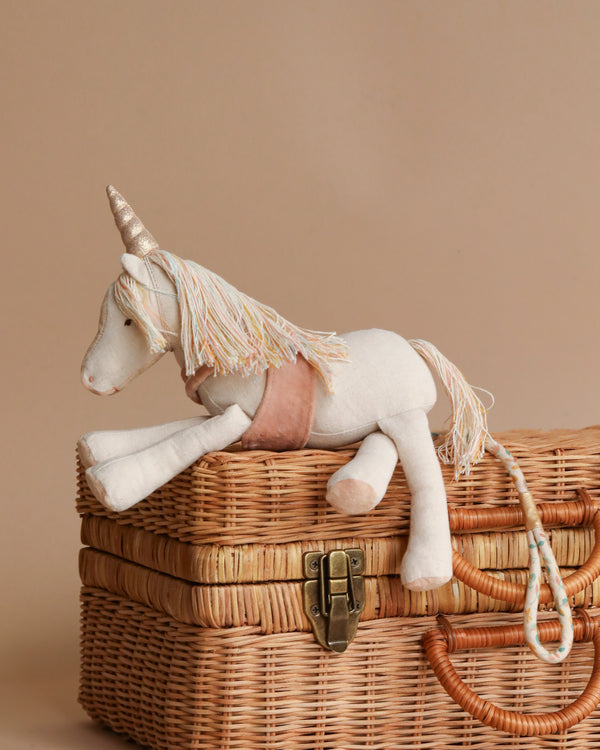 A Maileg Unicorn plush toy with a white body and a golden horn rests atop a woven wicker basket, featuring a soft beige background.