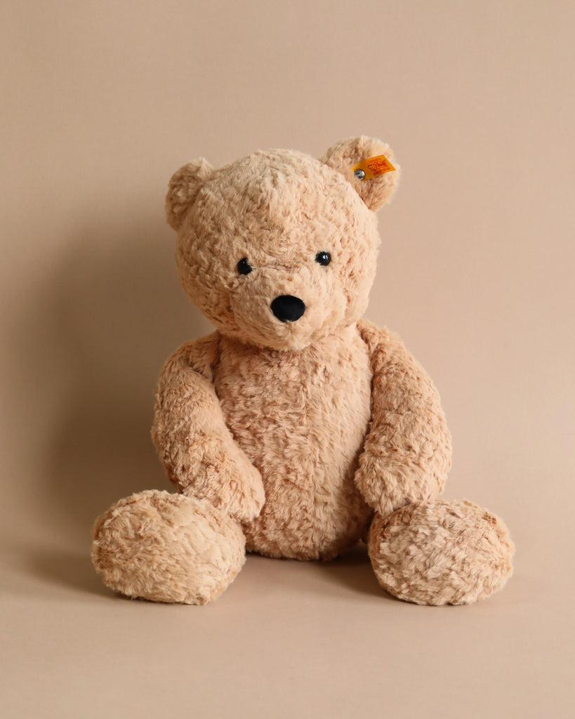 A Steiff Jimmy Teddy Bear, 16 Inches with a light brown, textured fur and a black nose, sits against a plain beige background. It has an alert expression with its head slightly tilted to one side and sports the distinctive