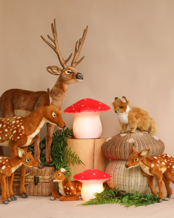 A whimsical display featuring a collection of animal figurines including a deer with antlers, several fawns, and a fox, arranged among large red mushroom lamps with an LED system and green foliage.