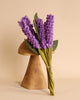 A Lavender Bouquet of handmade in Nepal felt lavender flowers with green stems, tied with twine, displayed in a wooden vase against a light beige background.