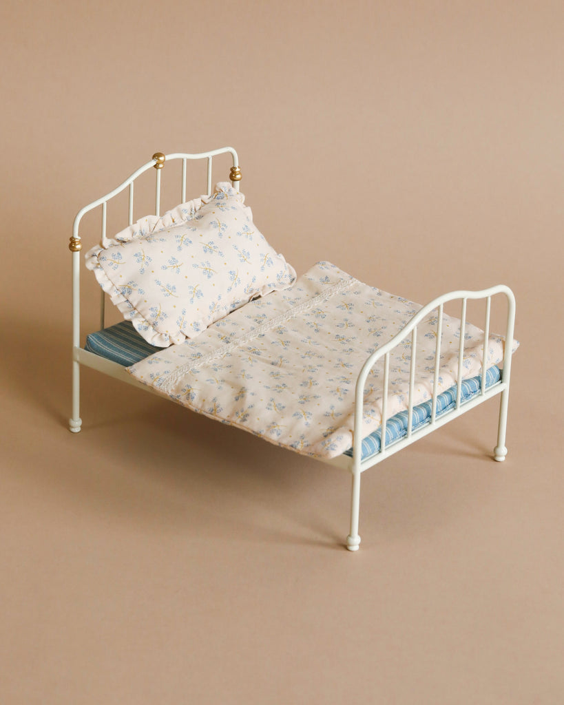 A Maileg Bed, Parent Mouse - Off White for bigger mice, adorned with Maileg printed fabrics on the pillow, blanket, and mattress in floral patterns. The bed features detailed railings and gold accents at the headboard and footboard. The background is a solid beige color.