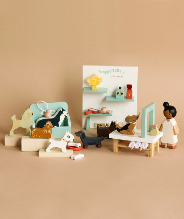 A wooden playset depicting a dog grooming salon, perfect for imaginative playtime. The Waggy Tails Dog Salon includes various wooden dog figures, representing different dog breeds, grooming accessories, and a small figure of a person. Grooming stations, a mirror, and dog-related items are arranged against a light brown background.