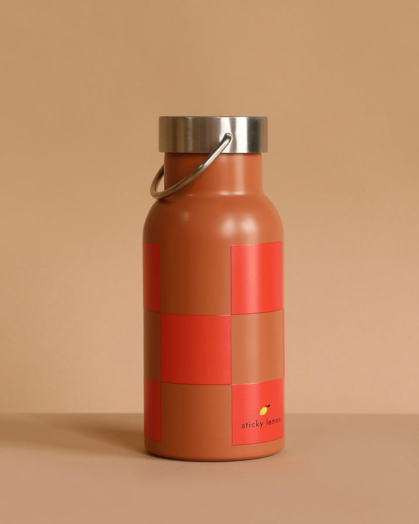 The Sticky Lemon Water Bottle | Farmhouse | Ladybird Red + Harvest Moon is a brown stainless steel bottle with a metallic lid and handle, featuring a red geometric pattern and small “sticky lemon” logo near the bottom on a beige background.