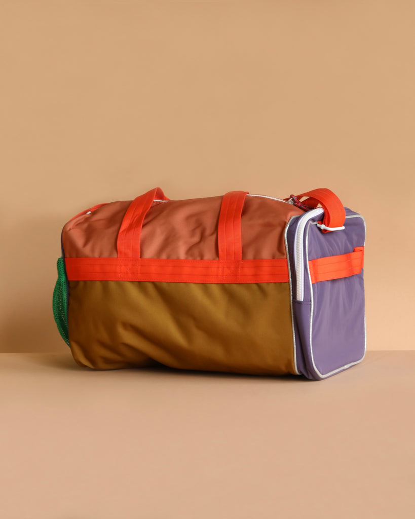 The Sticky Lemon Sports Bag | Better Together features orange handles, a brown front, purple sides, a green mesh pocket, and a tan top—all with waterproof lining and set against a beige background.
