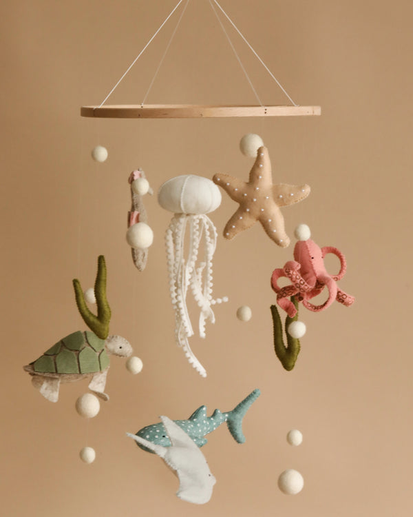 A charming nursery decoration, the Handmade Mobile - On The Reef - Final Sale features handmade felt marine creatures—turtle, jellyfish, starfish, octopus, and shark—handmade in Europe. Suspended from a wooden ring with small felt balls and seaweed accents against a plain beige background, it aids visual development for your little one.