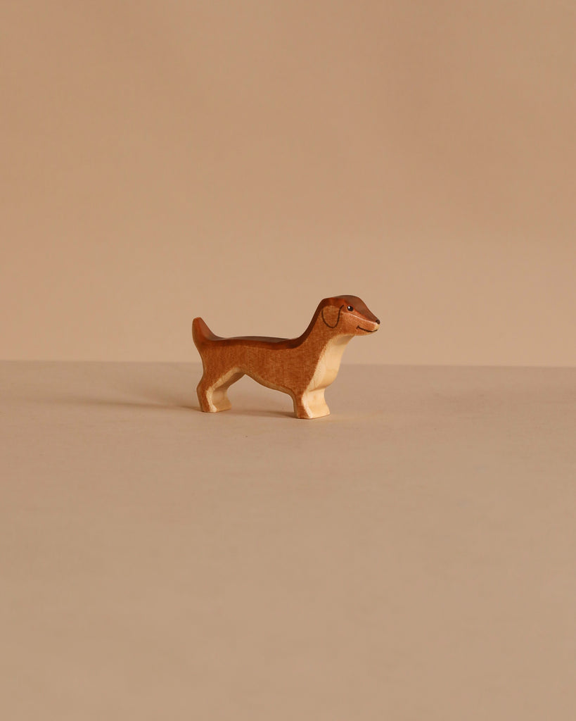The Handmade Holzwald Dachshund Dog, a small wooden figurine with a brown body and beige underside, stands on a plain beige surface with a matching background, highlighting the appeal of sustainable wooden toys.