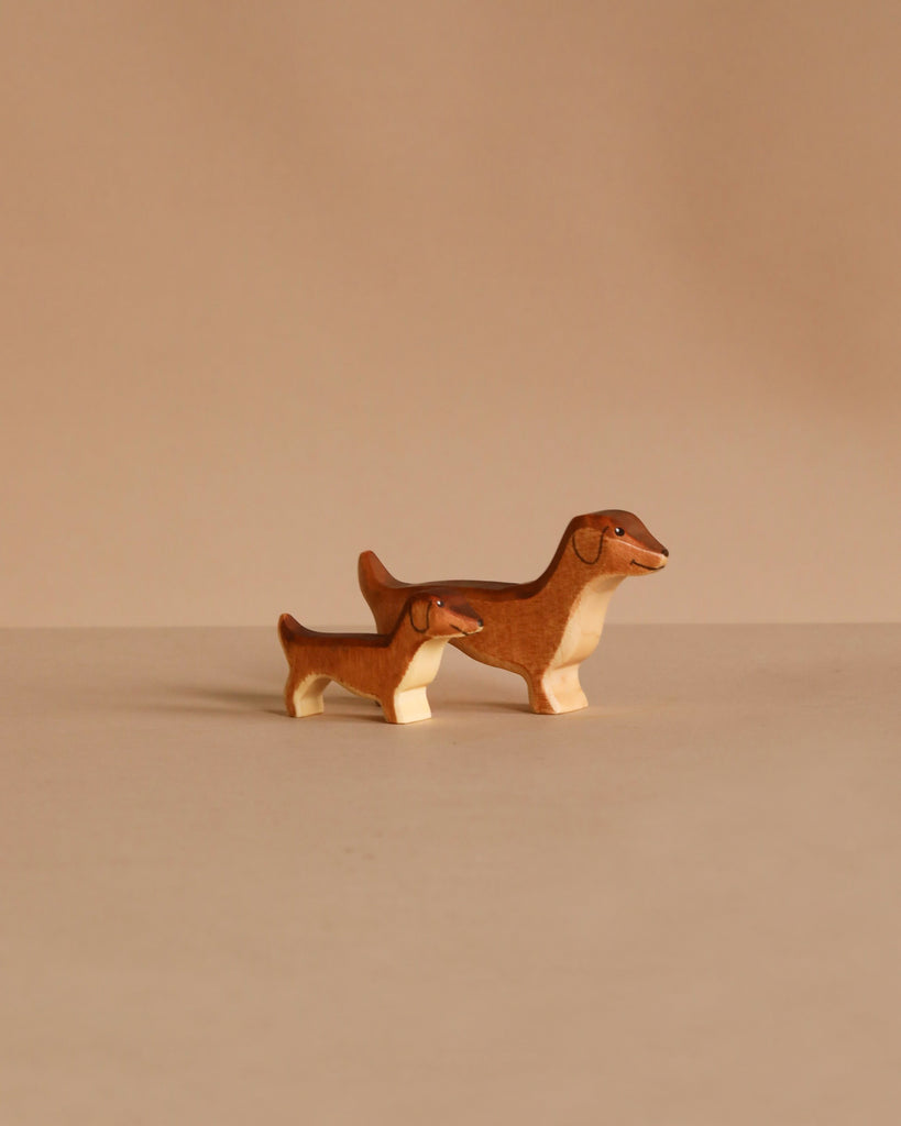Two small brown and cream Handmade Holzwald Dachshund Dog figurines stand side by side on a plain beige surface—charming, sustainable toys designed for mindful play.