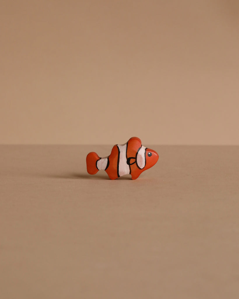 A Handmade Holzwald Clownfish, featuring orange and white colors with black stripes, sits on a plain beige surface with a matching background—an adorable addition to any Holzwald wooden toy collection.