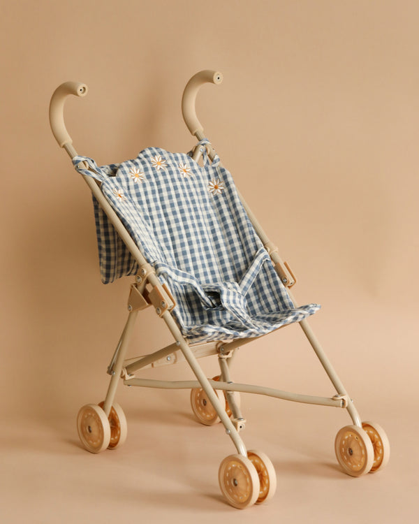 A lightweight beige children's stroller with double wheels and a blue and white checkered fabric seat, accented with small embroidered flowers. The Konges Sloejd Doll Stroller - Blue Checkered features curved handles and removable fabric, set against a neutral beige background.