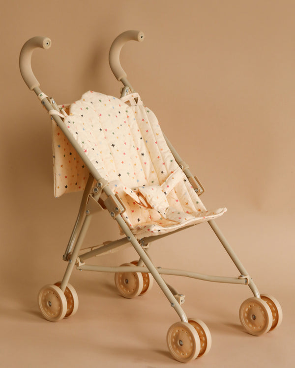 A beige folding stroller with curved handlebars and double wheels is set against a matching beige background. The Konges Sloejd Doll Stroller - Multi Star is adorned with a fabric cover featuring a pattern of small multicolored stars.