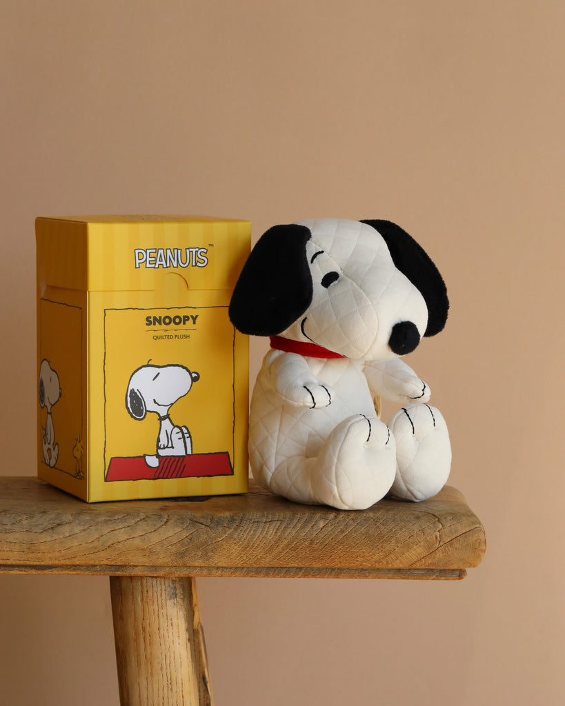 A Peanuts Snoopy Plush Toy in a gift box is placed on a wooden table against a plain beige background, with the box adorned by an illustration from the Peanuts comic strip.