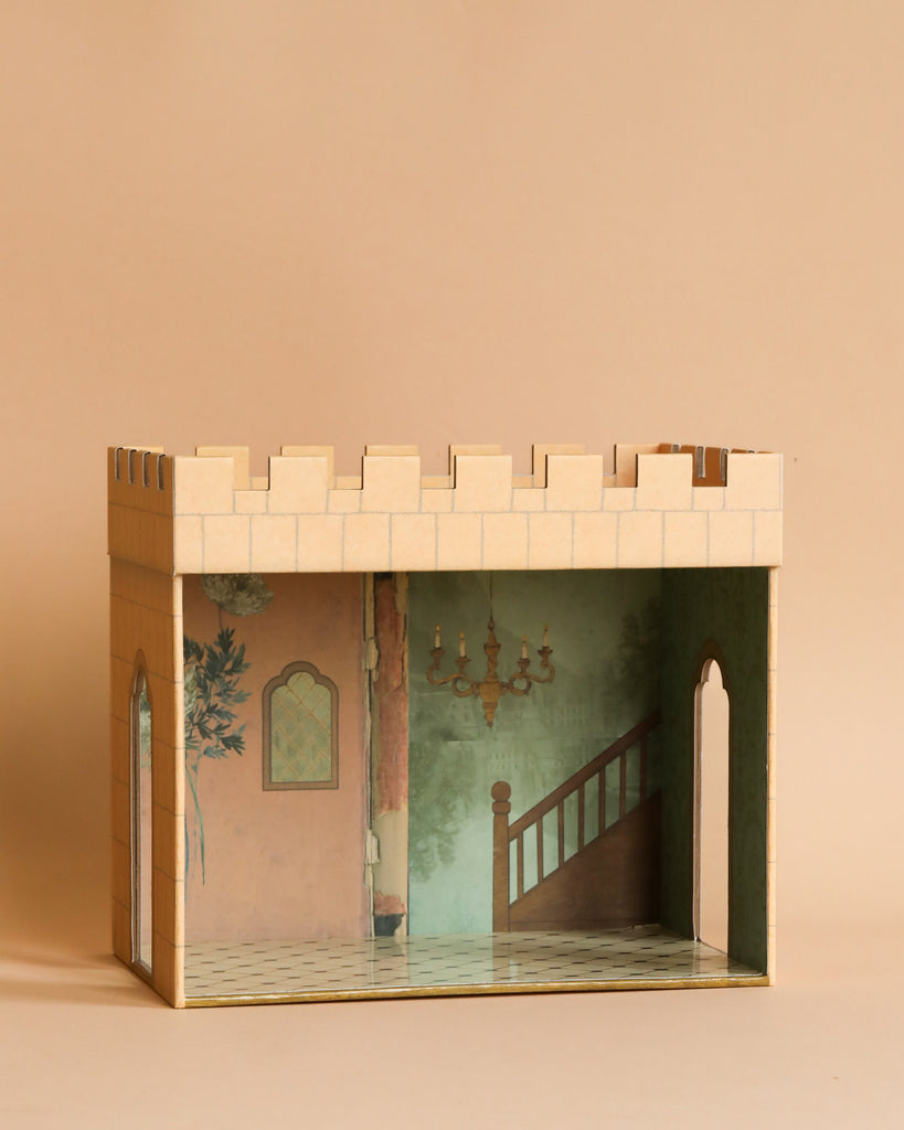 A small, open-roofed, cardboard playhouse resembling a Maileg Castle Hall. It has a beige exterior with crenellated walls and an interior featuring mural decor including a staircase, chandelier, and greenery. The structure stands on a plain, light brown background.