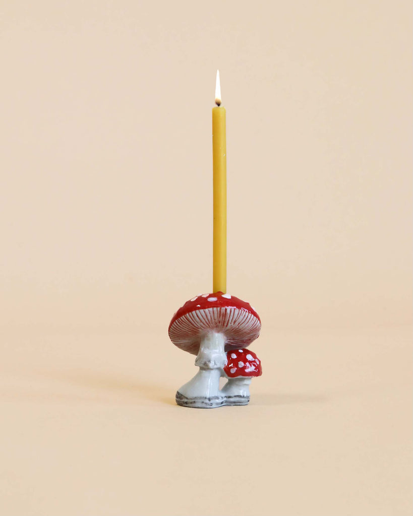A lit yellow candle standing in a whimsical, heirloom-quality Mushroom Cake Topper shaped like a red and white spotted mushroom against a pale beige background.