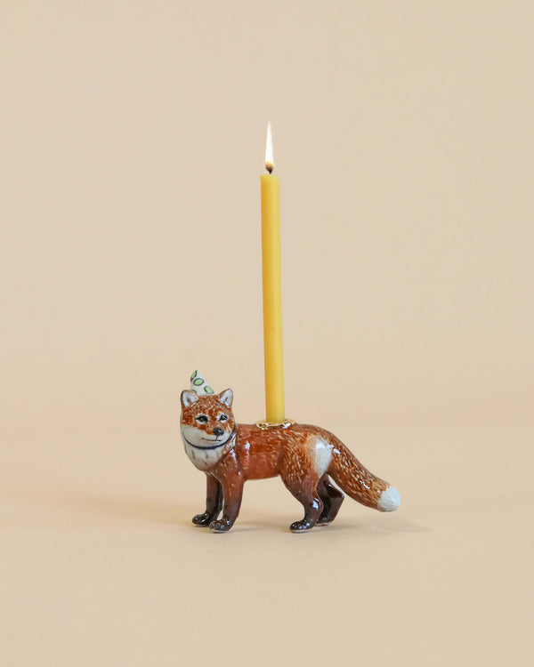 A whimsical Red Fox "Party Animal" Cake Topper shaped like a fox, with a lit yellow candle protruding from its back, staged against a plain beige background.
